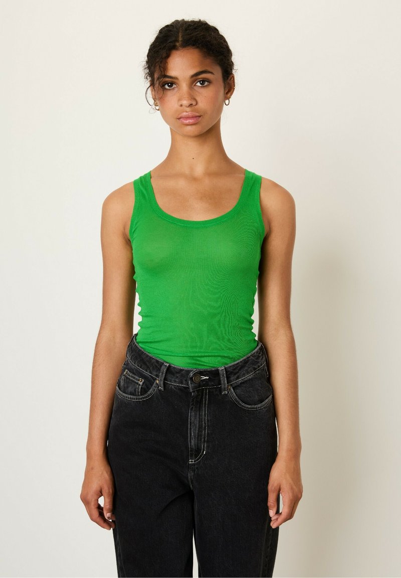 Žalias be rankovių topas su prigludusia forma, pagamintas iš lygaus audinio, derinamas su juodomis aukšto liemens džinsomis, turinčiomis priekines kišenes.