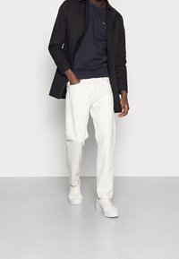 Zwarte jasje met knopen over een donkerblauwe trui, gecombineerd met witte spijkerbroeken en witte sneakers, met een ontspannen pasvorm en casual stijl.