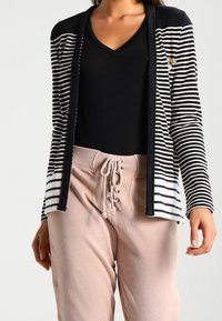 Cárdigan a rayas en blanco y negro con escote en V, que cuenta con un cuello negro liso y un pequeño emblema. Combinado con unos pantalones texturizados en rosa claro.