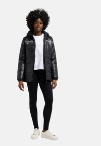 Svart pufferjacka med dragkedja fram, huva och elastiska ärmslut; har vit logotyp på bröstet; kombinerad med svarta leggings och vita sneakers.