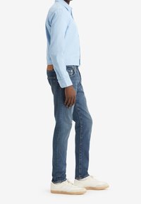 Camisa de manga larga azul claro, vaqueros azules de mezclilla y zapatillas blancas con suela texturizada, con un parche marrón en la parte trasera de la cintura.