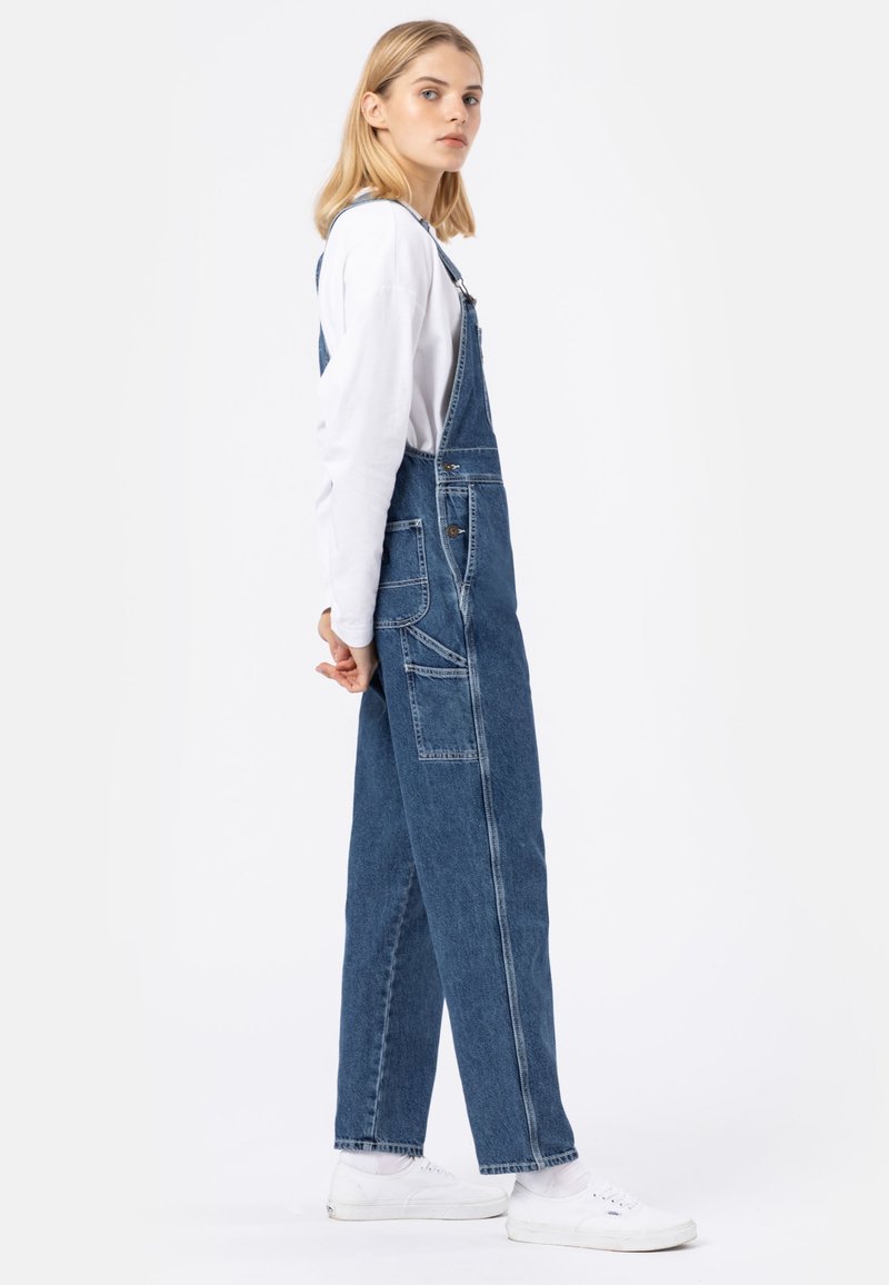 Salopette en denim bleue à coupe décontractée, avec des bretelles ajustables, des poches latérales et des ourlets retroussés, assortie à des baskets blanches.