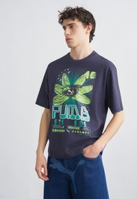 Νέος άνδρας που φοράει μπλε σκούρο φαρδύ T-shirt με μεγάλο πράσινο γραφικό λουλούδι και το κείμενο "PUMA", συνδυασμένο με μπλε τζιν, με το χέρι στην τσέπη.