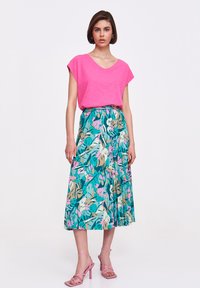 Różowa koszulka z dekoltem w serek w połączeniu z zwiewną, średniej długości spódnicą w tropikalny wzór liści w odcieniach teal, różu i beżu. Całość dopełniają różowe sandały na obcasie.