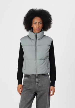 Grijze gewatteerde vest met hoge kraag, ritssluiting en zijzakken, gedragen over een zwart langemouwenshirt met een gestructureerde stof.