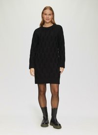 Schwarzes gestricktes Kleid mit einem strukturierten Muster und ovalen Ausschnitten, langen Ärmeln und einem runden Ausschnitt, kombiniert mit schwarzen klobigen Stiefeln.