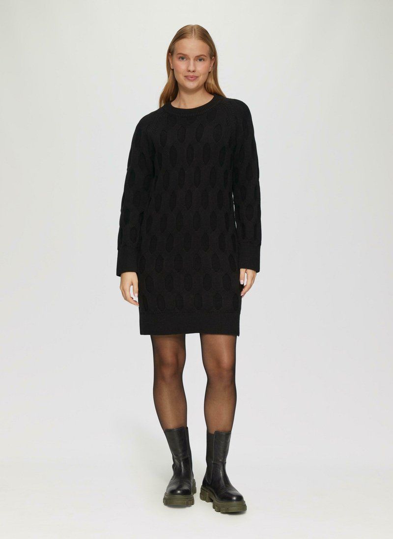 Schwarzes gestricktes Kleid mit einem strukturierten Muster und ovalen Ausschnitten, langen Ärmeln und einem runden Ausschnitt, kombiniert mit schwarzen klobigen Stiefeln.
