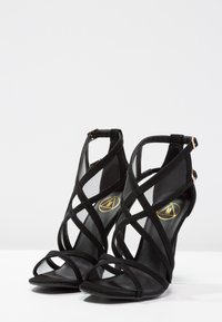 Korkeastaiset mustat sandaalit, joissa on risteävät verkkokankaiset ja mokkasiinistrapet, avonainen kärki ja tyylikäs korko kultaisella logolla sisäpohjassa.