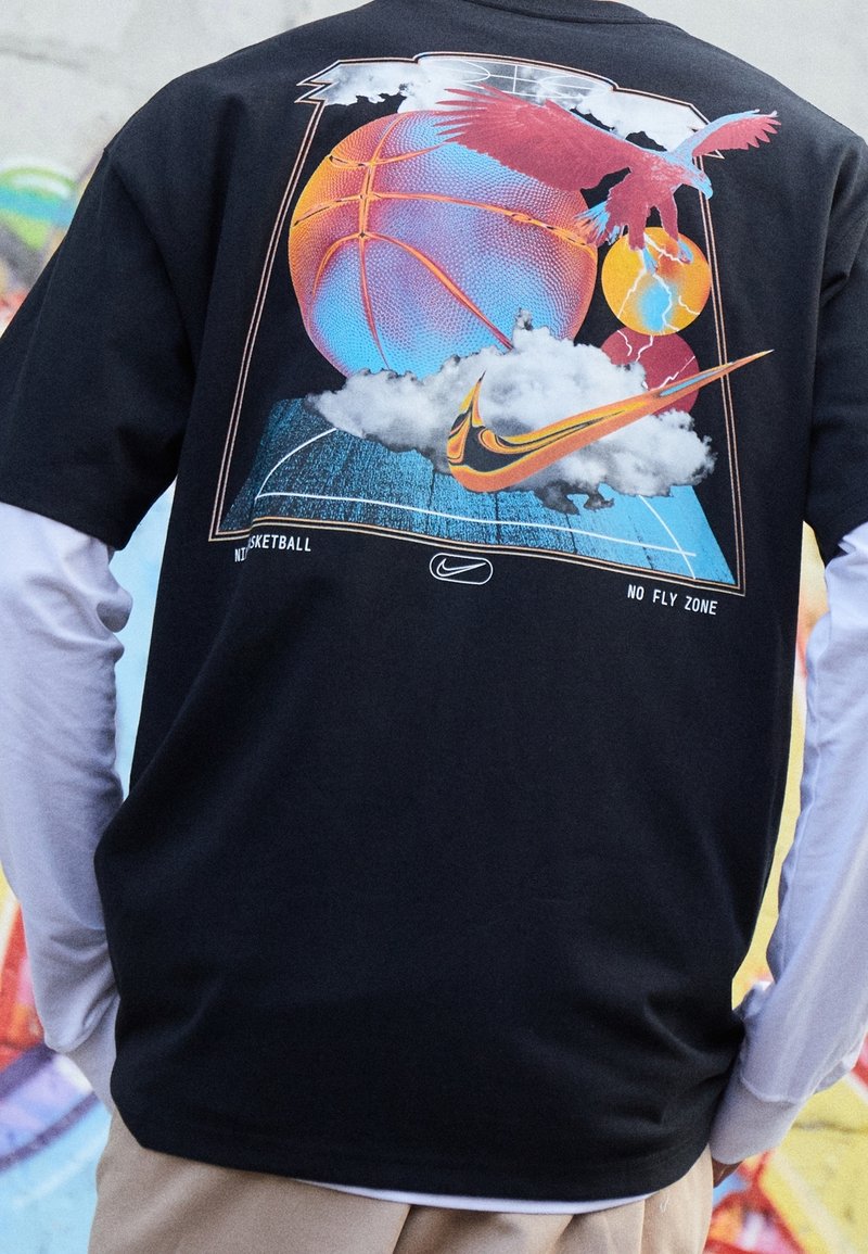T-shirt noir avec un design graphique coloré incluant un ballon de basket, des nuages et un aigle, ainsi que le logo Nike et le texte "NO FLY ZONE".