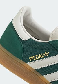 Zelené semišové a bílé kožené tenisky se třemi bílými pruhy, zlatým textem "SPEZIAL" a gumovou podešví. Viditelné texturované detaily na svršku.