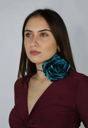 Giovane donna con lunghi capelli castani che indossa una maglia bordeaux e una collana rigida color teal a forma di rosa, guarda leggermente verso l'alto e di lato.