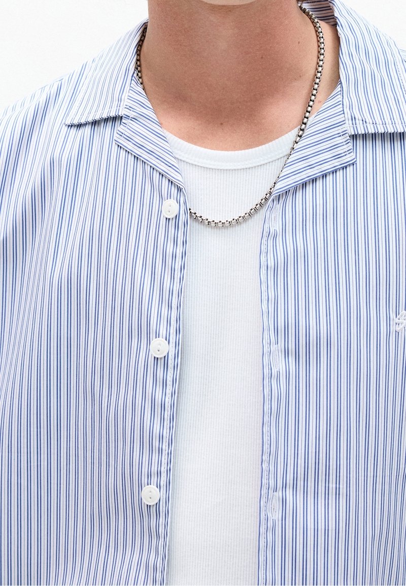 Personne portant un collier chaîne argenté, un t-shirt blanc côtelé et une chemise ouverte rayée bleu et blanc avec des boutons.