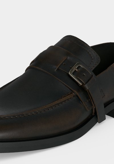 Gros plan d’un mocassin en cuir brun foncé avec une bride et un détail de boucle argentée sur un fond clair.