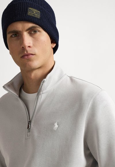 Νέος άνδρας που φοράει ένα σκούρο μπλε πλεκτό σκουφάκι και ανοιχτό γκρι πλεκτή μπλούζα με φερμουάρ μέχρι τη μέση, με ένα λευκό κεντητό λογότυπο αλόγου στο στήθος.