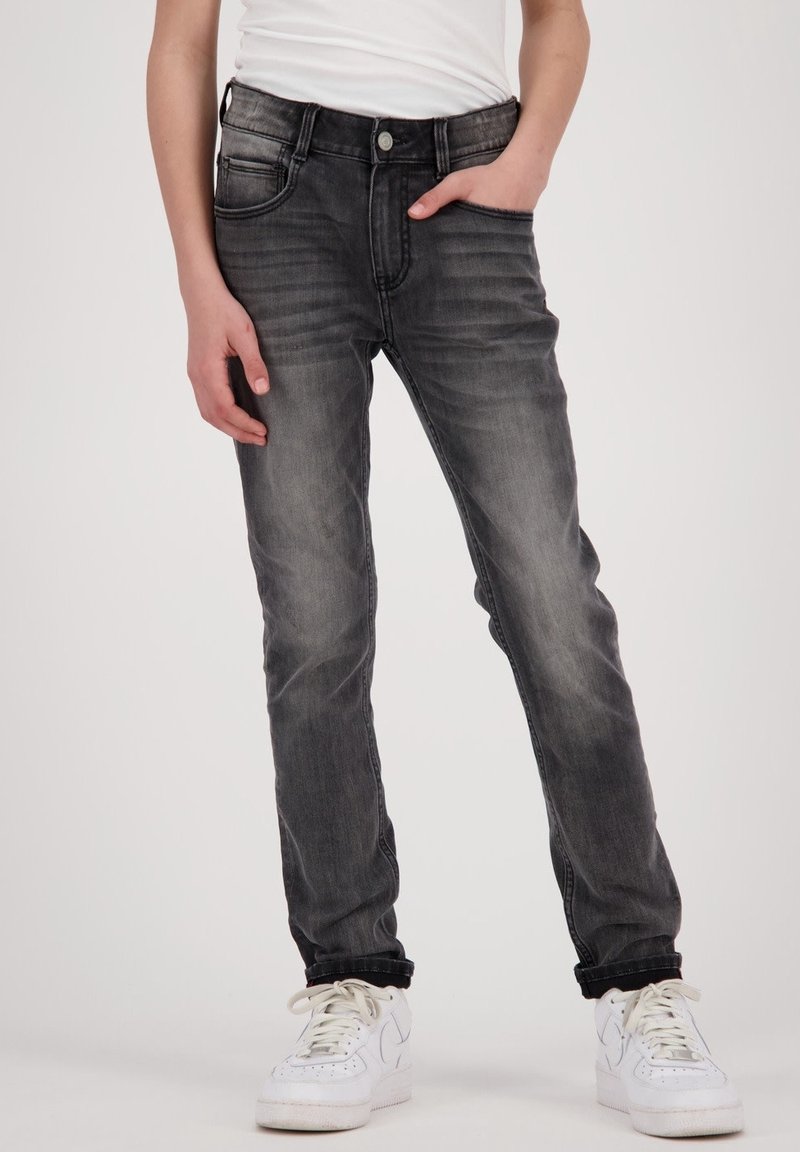 Jean slim gris foncé avec un effet délavé, design à cinq poches et légère usure aux plis. Porté avec une chemise blanche unie et des baskets blanches.