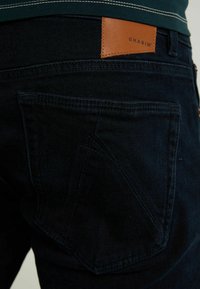 Vaqueros de mezclilla azul oscuro con un bolsillo en ángulo distintivo, parche de cuero marrón de la marca y detalles de costura sutiles.