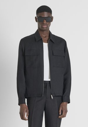Zwarte zip-up jack met twee voorzakken, gemaakt van een gestructureerde stof, gecombineerd met een witte geribde tanktop en donkere broek. Zwarte zonnebril gedragen.