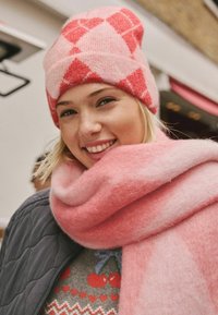 Rosa und rote gemusterte Strickmütze und passender rosa Schal, beide aus weichem, strukturiertem Stoff, getragen über einem grauen Pullover mit dekorativen Akzenten.