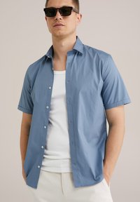 Camisa de manga corta de botón en tela azul claro, que presenta un cuello, siete botones y un corte relajado, llevada sobre una camiseta sin mangas blanca.