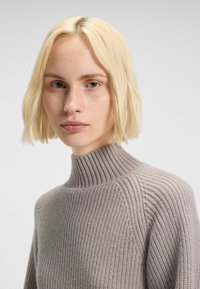 Young person with short blonde hair wearing a light gray rib-knit turtleneck sweater, facing forward against a plain background.