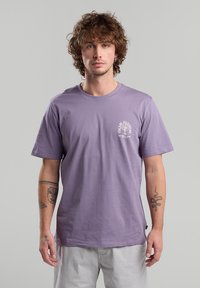 Short-sleeve purple cotton t-shirt featuring a white graphic design on the left chest and a relaxed fit. Model wearing light-colored shorts.