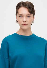 Felpa teal con collo a giro, morbida al tatto e dalla vestibilità rilassata. Presenta cuciture sottili e maniche con spalle scese.