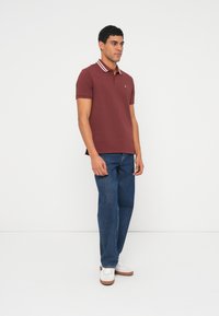 Álló férfi, bordó galléros pólót visel, amelyen fehér csíkok díszítik a gallért, kék farmernadrágot és fehér tornacipőt, egyszerű fehér háttér előtt.