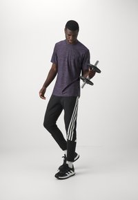 Figura maschile che tiene un manubrio, indossando una maglietta atletica viola con texture e pantaloni da jogging neri con strisce bianche. Scarpe da ginnastica nere.