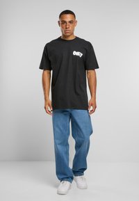 Melnas kokvilnas T-krekls ar baltu "OBEY" logo, brīva piegriezuma, kombinēts ar gaiši zilām denima biksēm un baltām sporta apaviem, uz viendabīga fona.