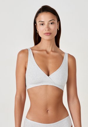 Femme aux cheveux raides et foncés portant un bralette cache-cœur sans manches gris clair et un short assorti sur un fond uni.