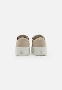 Beige Canvas-Slipper mit strukturierter Oberseite und weißer Gummisohle. Verfügt über ein kleines Logo an der Ferse und ein niedriges Profil.