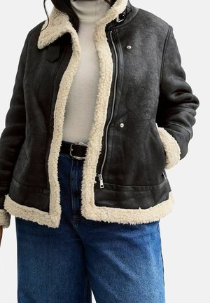 Czarna kurtka z owczej skóry z kremowym futerkiem, noszona na białym golfie i niebieskich dżinsach, ręka w kieszeni.