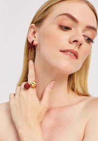 Czerwone kolczyki w kształcie kwiatów z żywicy i pierścionek w czerwono-złotym kolorze nosi osoba o gładkiej skórze i prostych blond włosach na jasnym tle.