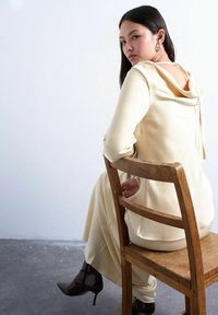 Femme en robe crème à dos nu assise de côté sur une chaise en bois, portant des bottines à talons brun foncé et des boucles d'oreilles créoles en or.