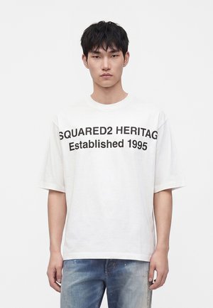 Mladý muž s krátkymi čiernymi vlasmi, ktorý nosí biele tričko s čiernym textom "SQUARED2 HERITAG Established 1995" a vyblednuté modré džínsy.