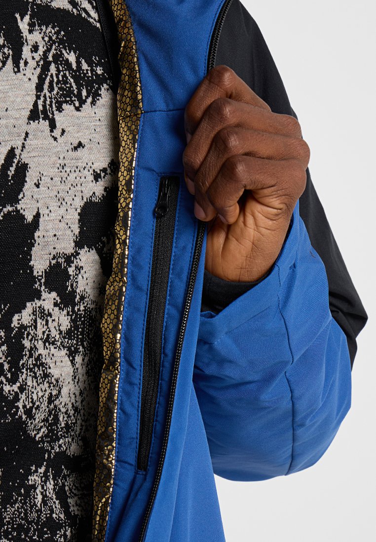 Blaue Jacke mit schwarzen Ärmeln, ausgestattet mit einer Taschen mit goldfarbenem Reißverschluss. Strukturieret Innenfutter sorgt für zusätzlichen Detail. Trägt darüber ein gemustertes Hemd.