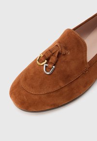 Brown suede loafers featuring a rounded toe and a decorative lacing with gold and silver hardware accents. Soft texture with minimalist design.