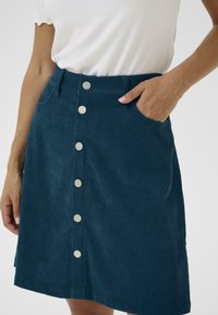 Teal kordonhame, jossa on nappikiinnitys edessä, sivutaskut ja pystyryppyinen pinta. Hameessa on istuva vyötärö ja levenevä helma.