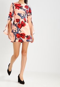 Blomsterkjole i fersken med røde og blå mønstre, med klokkelængde ærmer og en tætsiddende talje. Båret med sorte høje hæle.