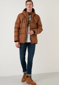 Brun oppustelig jakke med hætte, med frontlynlåse, sorte detaljer og kviltet design. Bæres med blå jeans og lysebrune støvler.