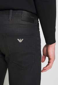 Svart denimjeans med en slät yta, försedd med en läderetikett från Emporio Armani och en metallisk logotyp på bakfickan.