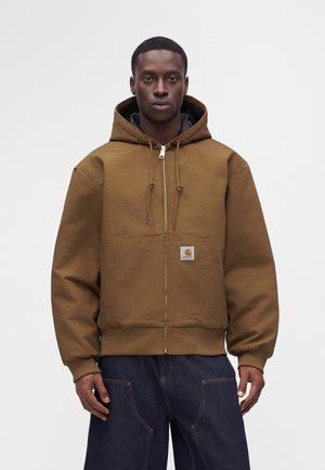 Homme à la peau foncée portant une veste à capuche marron zippée avec une poche avant et un pantalon bleu foncé, debout devant un fond clair uni.