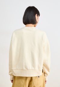 Sweatshirt couleur crème avec une coupe décontractée, épaules tombantes et ourlet côtelé. Porté avec un pantalon marron clair, avec des coutures visibles.