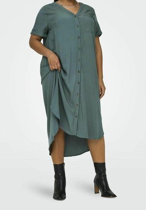 Personne portant une robe midi verte boutonnée avec des manches courtes et des bottines noires à talons, soulevant légèrement le côté de la robe.