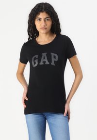 Zwarte katoenen T-shirt met een ronde halslijn en korte mouwen. "GAP" logo in een vet, grijs lettertype centraal op de voorkant geplaatst.