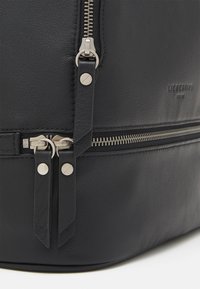 Sac à dos en cuir noir avec des zips et des accessoires en argent, rabat repliable, et surface texturée avec des détails de couture visibles.