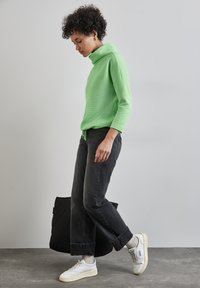 Grüner strukturierter Rollkragenpullover, schwarze weite Jeans, weiße Sneakers, trägt eine schwarze gesteppt Tasche, steht vor einem neutralen Hintergrund.