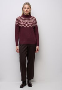 Vínový pletený svetr s krémovým zigzag vzorem na výstřihu, doplněný tmavě hnědými kalhotami a hnědými kotníkovými botami.