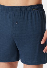 Pantaloni da bagno blu navy con un motivo a griglia testurizzato, cintura elastica e un dettaglio a bottone sul davanti. Realizzati in tessuto leggero.