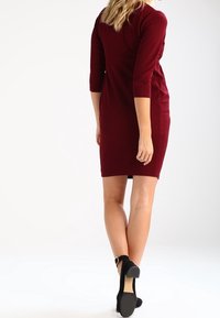 Vinröd knälängd klänning med trekvartslånga ärmar, figurnära passform, ribbad textur och en slät tygfinish. Bärs med svarta högklackade skor.